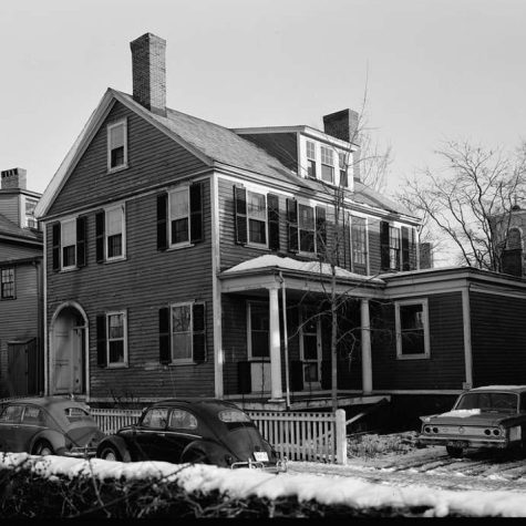A Cambridge multifamily building sits swaddled by snow. On the street are two Volkswagen beetles. Chimneys stick out of either end of the roof.
