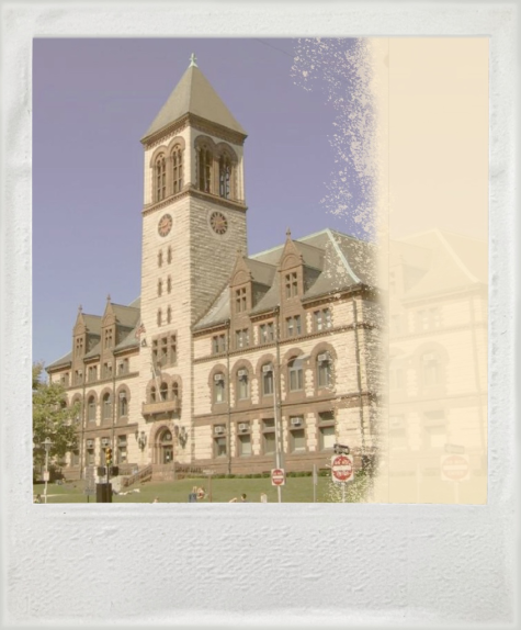 A photograph of City Hall in Cambridge, Massachusetts. It is a multi-story stone building with a tall obelisk-shaped central tower.